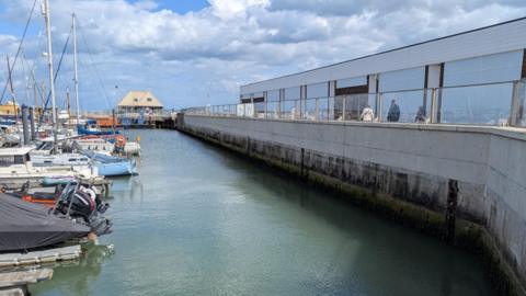 A pier with boats parked in the harbour. The harbour is enclosed within a stone wall and glass panelling running on top. 
