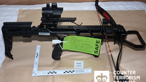 A black crossbow with a right-angle ruler beneath and a green police label attached reading "Safe" and other details that cannot be made out. The weapon is sitting on light brown paper