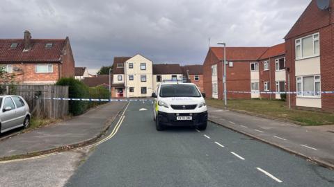 A road with a police van parked facing towards the camera. A blue and white police cordon is closing off the area. Houses are surrounding the road. 