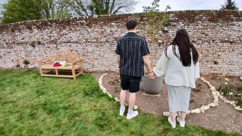 A man and a woman stood next to each other holding hands and looking down at a small tree in a pot which is flanked by white rocks in a green garden. There is a bench with bunches of flowers next to them.