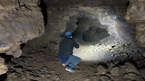 A man kneeling down, shining a light further down the tunnel.