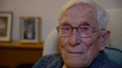 A close up shot of John Daniels at the age of 104. He has neat, white hair, and gold framed glasses. He is wearing a hearing aid, and a thick, knitted blue cardigan. He is sitting in a high backed armchair in his living room. Behind him on a dresser and out of focus, is a framed photo of him as a young man in Navy uniform.