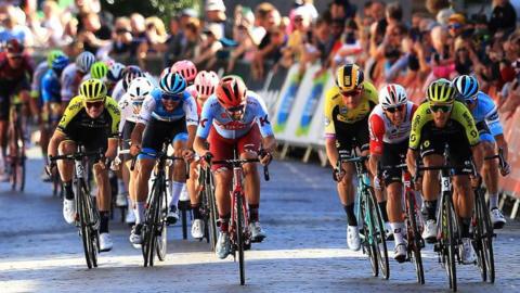 A peloton of male cyclists racing full pelt on a cobbled street. They are all wearing brightly coloured lycra and helmets