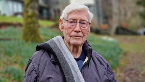 Alan Kelly outside Hawes End. He has short white hair and blue eyes, with glasses with a thin metal frame. He is wearing a grey coat and has a striped grey scarf over one shoulder. He is looking at the camera with a serious expression. Behind him is Hawes End, a former orphanage, in the blurred background.