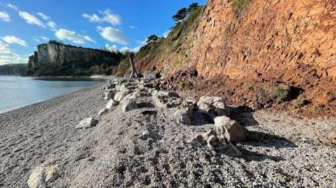 A picture of the seafront. There is a number of rocks on the floor in front of the cliffs. The sea can also be seen to the left of the image. 
