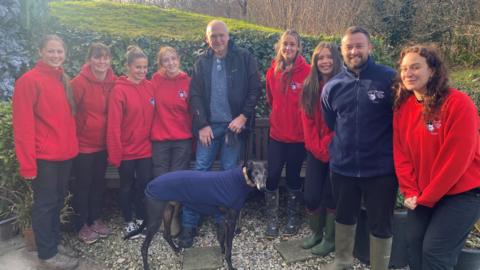 Nine people stood together outside with the greyhound dog, Bobby, at the centre. The dog is wearing a blue coat. The volunteers, stood either side of Peter Hart, are wearing red coats. Peter is wearing a black coat. They are all smiling at the camera.