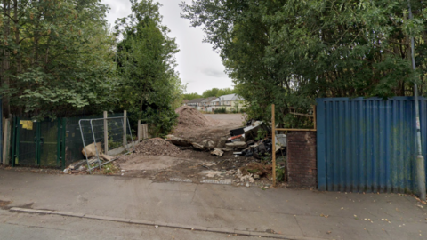 Trees and bushes are to the left and right of an entrance. Part of a road and pavement are at the bottom of the photo. 