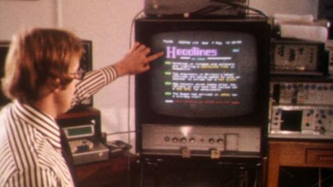 A man points at a television screen where Ceefax is on display. He sits beside a range of technical equipment.