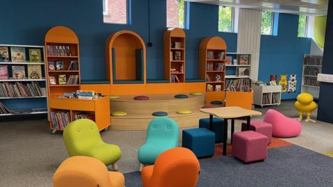 A brightly coloured children's section of a library in Staffordshire. There is chunky furniture in various colours, and orange bookshelves set against blue walls. There is a stepped seating area with coloured cushions.