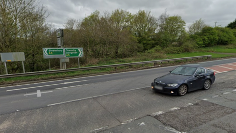 A road junction with a dual carriageway in front of it. A black BMW is travelling along the road. The other carriageway is clear. There are green signs behind a crash barrier, the one on the left reads "Shrewsbury A5" and the one on the right reads "Oswestry, Llangollen A5".