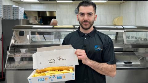 Chris Onisiforou is pictured standing in a fish and chip shop. He is holding a portion of fish and chips in an box open. He is wearing a black polo shirt and he has short black hair which is gelled back and a short beard. 