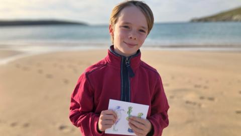 A young boy with blond hair and a red fleece stands of a beach in the sunshine, hoping an illustrated greetings card 