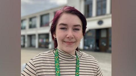 Ms Reilly has short purple hair and is sitting, smiling at the camera. She's wearing a striped top and a sunflower lanyard.