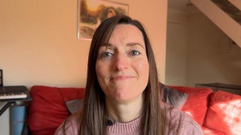 Natasha has long brown hair and is wearing a lilac top and sitting on a red sofa. 