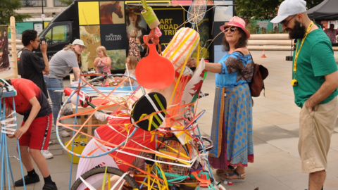 A quirky art piece made up of colourful objects such as tennis rackets and hoola hoops, pieced together. Two people are admiring the piece, a woman in a long blue dress with sunglasses and a pink hat and a man in a green top and beige shorts with a grey baseball cap. The art piece is outside, other activities are taking place in the background.