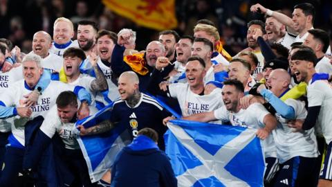 The Scotland's men's football team gather together for a group photo after winning their match against Denmark. They are wearing tops which say "We'll be coming" and holding blue and white Scotland flags. Behind them, fans can be seen in the stands waving a yellow and red Lion Rampant flag.