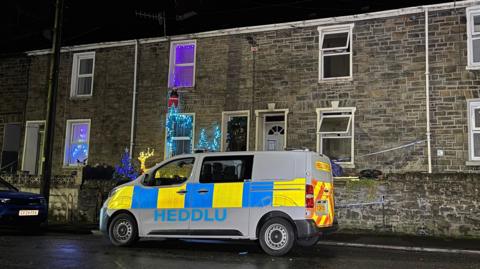 Photo of a police van outside a line of houses. One of them has lots of Christmas lights up on the wall. The next house has all its windows open