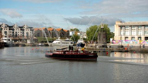 A black boat with red stripes sailing in Bristol Harbour. There are buildings in the distance.