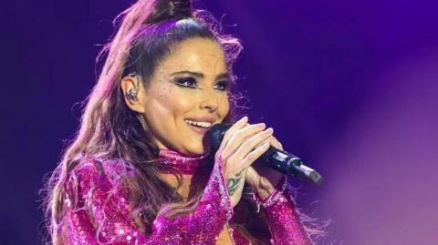 Cheryl singing into a microphone with a purple background. She wears a sequined pink costume and has sequins stuck on her cheek. There is an earpiece in her ear and her hair is half up in a knot on top of her head with the rest cascading down past her shoulders