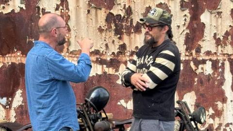 Jason Momoa standing talking to an Electrogenic staff member. Next to them is a vintage motorcycle.