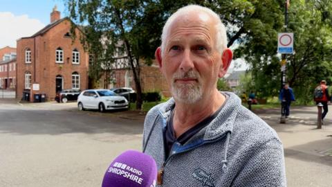 A man with grey hair and grey beard interviewed in a street. He's wearing a grey top