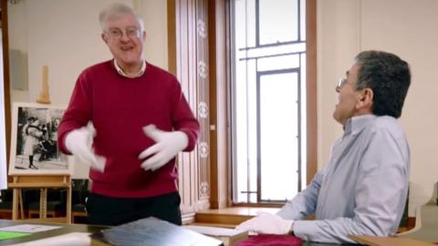 Mark Drakeford looking happy mid dance. He is wearing a red jumper and white gloves. Next to him is a man looking up with the same type of white gloves. They're in a brightly lit room with a picture on an easel behind them.