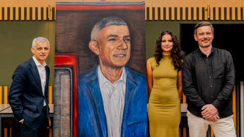 Sadiq Khan, Tia Mirza and Jonathan Carr pose next to the large portrait of the mayor. Khan, who has short white hair, is wearing a dark blue suit and a white shirt with an open collar. Mirza has long dark curly hair and is wearing a mustard-coloured dress with short sleeves. Carr has short brown hair and a goatee beard and is wearing white trousers and an open-collar black shirt.