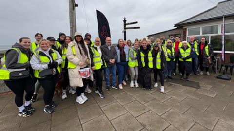 A group of about 30 people, men and women, and a mix of ages but mainly in their early 20s about to set off on a charity walk. Some are wearing hi-viz jackets and one person at the front is carrying a collection bucket. They are standing on a pavement, with a cafe building to the right, and the sea can be seen behind them.