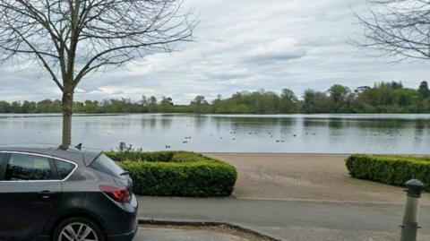 A black car is parked in front of a pathway with a large lake with ducks swimming.