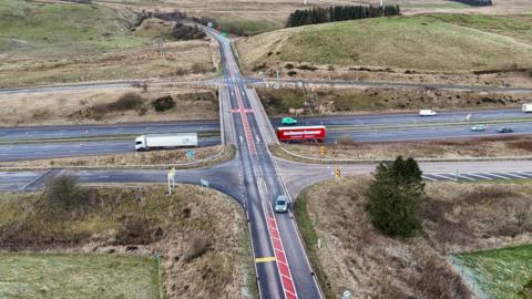 Aerial view of a junction - the A684 crossing the M6 in Cumbria. The A road features red hatching between the carriageways. Lorries and cars are being driven along the motorway which is in an area of scrubbly grass and a few trees.