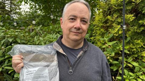 A man in a grey quarter-zip holding a crumpled package from Royal Mail with an apology letter, stood in front of a green bush.