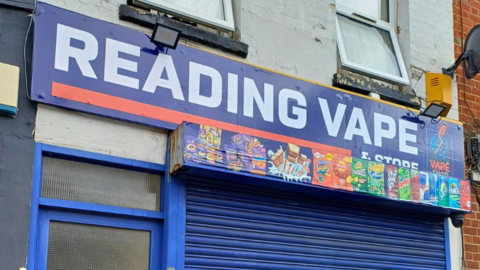 A general view of Reading Vape, which is has blue shutters, a blue door on the left of the picture and a blue sign with white lettering that spells out "Reading Vape and Store". 