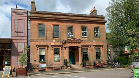 An old sandstone building in Dumfries with bicycles parked outside and a tree at its side