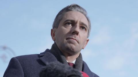 Simon Harris addressing reporters in Dublin on 20 November.  He is wearing a dark grey coat, a grey scarf, a light shirt and a maroon tie. 