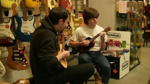 Two young men with dark hair sit on stools while playing the ukelele on the shop floor. There are guitars of various colours dispalyed on the wall behind them.