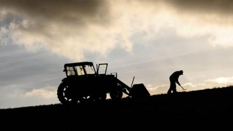 A tractor and farmer raking the ground are silhouetted against a blue sky with yellow clouds.