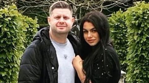 Josh and Chloe Bashford embrace looking towards the camera in a garden. On the left Josh has short light brown hair and a stubble beard. He wears a grey t-shirt and a black puffer coat. On the right Chloe has long black hair and wears a black jumper with diamante on the sleeve. 