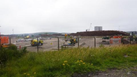 A site at the Port of Workington, which is being used for the development of a facility to support the decommissioning of a nuclear reactor in Japan. The site is fenced off and there are heavy plant machines on the concreted yard. In the distance there are cranes and large tanks.