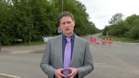 Nicky Schiller, wearing a suit and tie, reports in front of a police cordon on a road near the track