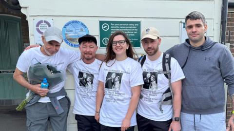 Josh Bashford's father, two brothers, his sister and her partner stand together in front of the Vanguard Way finish sign. They wear while t-shirts with a black and white photo of Josh and Chloe.