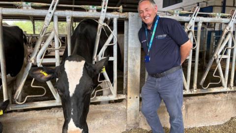 Philip Steans stands on the right, smiling at the camera. He wears a blue polo top and trousers and has a lanyard on. He is standing in a barn beside a cow, which has its head through a metal feeder