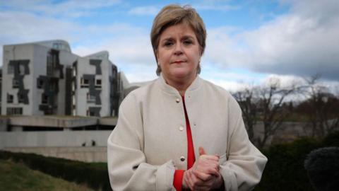 Nicola Sturgeon in white jacket and red shirt is standing rubbing her hands outside the Scottish parliament