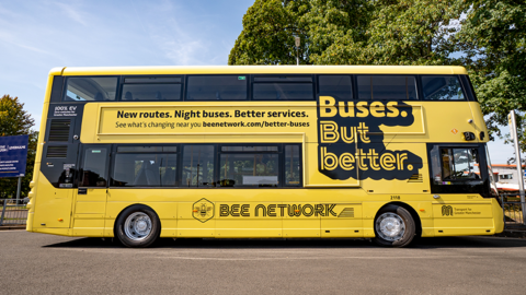 An image on the side on of a stationary yellow double-decker bus, with trees in the background.