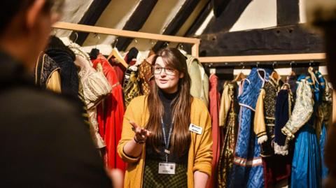 A woman with long brown hair and glasses is wearing a mustard yellow cardigan, black top and yellow and black skirt. she is standing i front of two racks of Tudor costumes