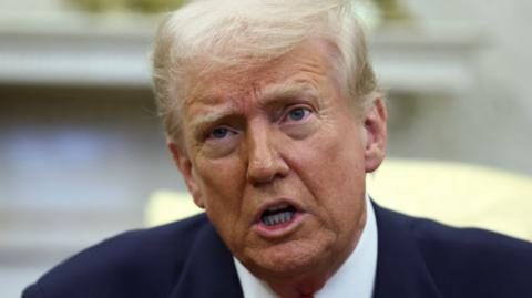 Donald Trump, wearing a blue suit and red tie, looks up as he speaks to reporters in the Oval Office