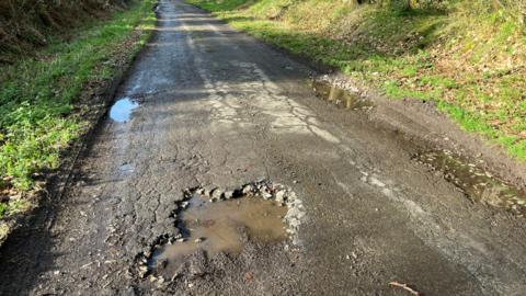 A narrow country road with grass verges on either side and a large pothole filled with water in the foreground