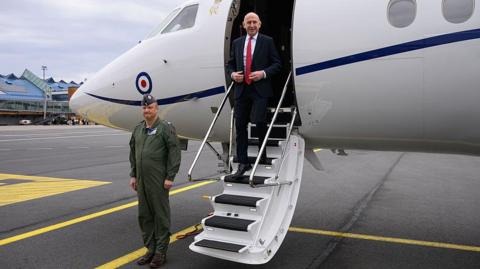 John Healey in a white shirt, black suit and red tie, seen exiting an RAF plane at an airport as a soldier stands to attention nearby
