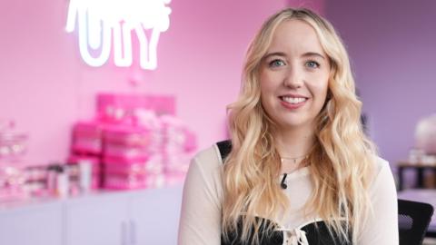 A woman with long blonde hair in a black and beige shop, with a pink-purple blurred background