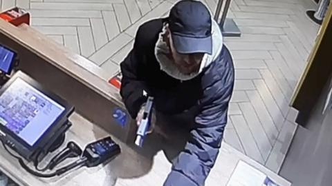 A man is holding a gun at a counter, he is wearing a black coat and cap