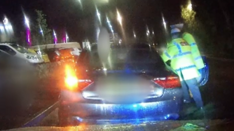 The dashcam footage shows a night-time scene. Two police officers, wearing high visibility jackets, are at the open driver's door of a blue car.